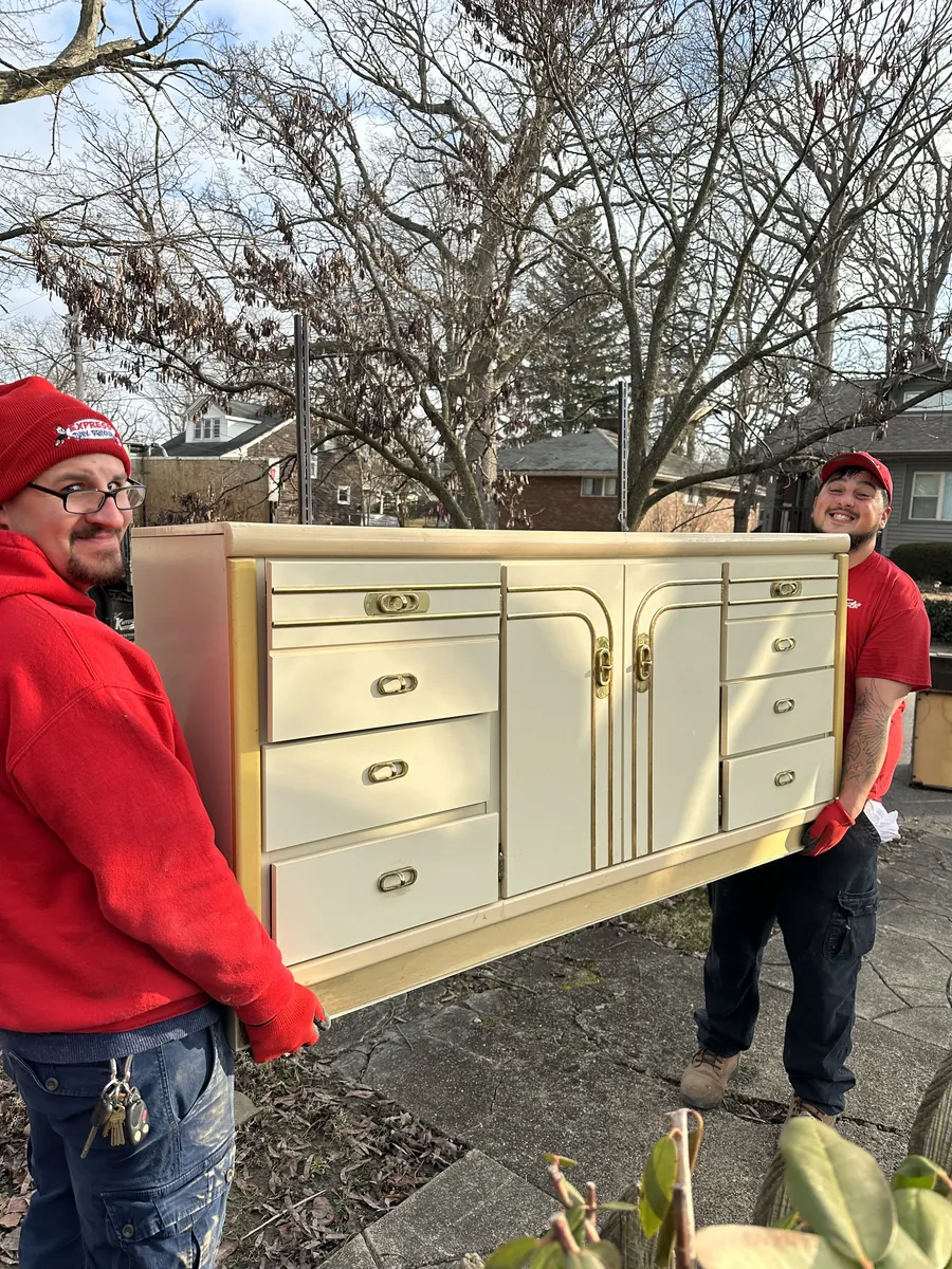 Two Express Junk Removal crew members carrying a dresser out of a home